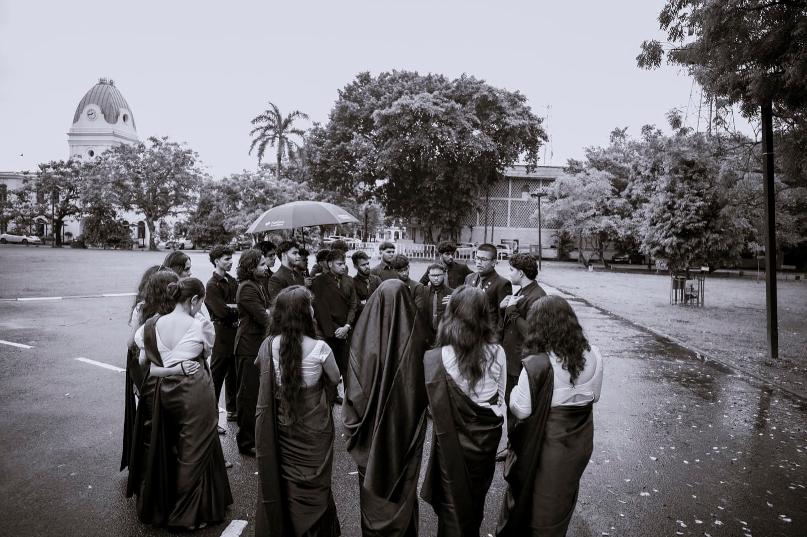 Club members in sarees and suits gathered on a wet road, one holding an umbrella